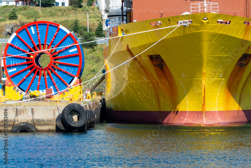 Bulk sub-sea industrial glass fiber optic cable on a metal spool on a ship's stand. The yellow data line is coiled around a black reel in a storage yard. Internet communications spool storage yard.