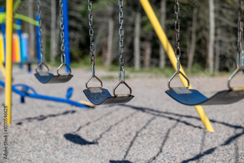 There are three school swings with black rubber seats hanging from metal chains. The children's swing set has yellow metal legs. The ground is covered with small pebbles. A green fo in the background.