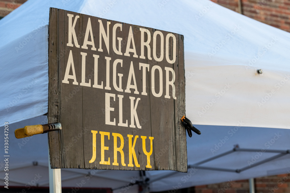 Fototapeta premium A large black outdoor sign with kangaroo, alligator, elk, and jerky in white and yellow text. The meat variety is for sale. The signage is on a pole next to a white tent at a farmer's market. 
