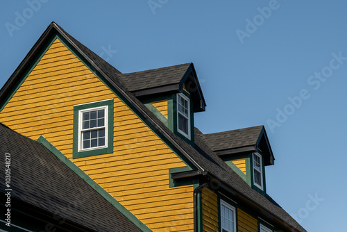 The top exterior floor of a yellow colored cape cod house with green and white trim. The building has two peaked dormer windows. The roof is covered in black asphalt shingles. The sky is bright blue.