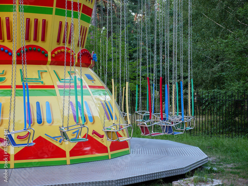Empty old-fashioned merry-go-round in a city park. Red and blue wooden seats on metal chains of swing carousel. Trees on the background, fallen leaves on the ground