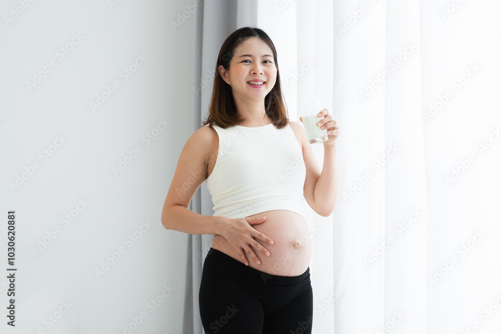 Pregnant Asian young woman standing and holding a glass of fresh milk near window at home and touching her belly with love and care. Pregnancy lifestyle. Happy mom smiling at camera Pregnant Asian young woman standing and holding a glass of fresh milk near window at home and touching her belly with love and care. Pregnancy lifestyle. Happy mom smiling at camera