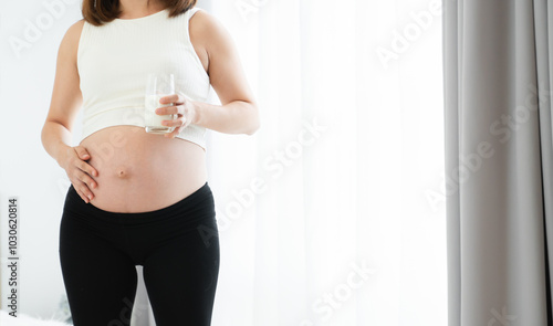 Pregnant Asian young woman standing and holding a glass of fresh milk near her belly with love and care. Pregnancy lifestyle. Copy space. Selective focus on hand and milk