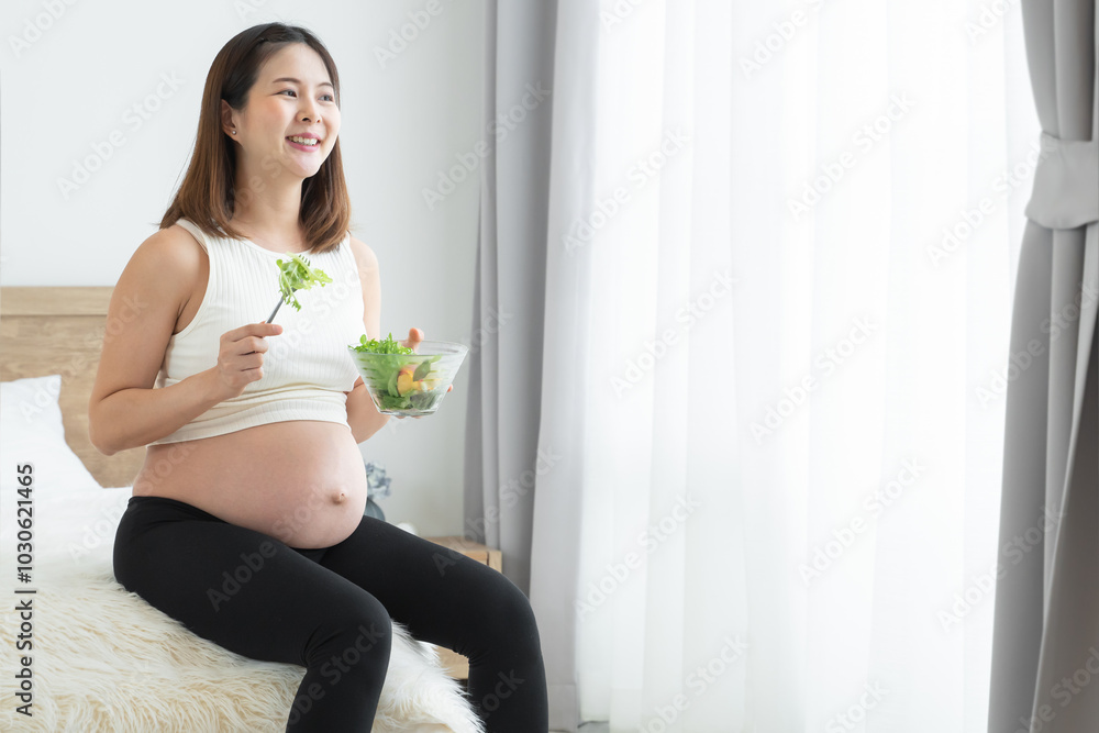 Young pregnant woman is sitting, holding a bowl of salad and fork. She is smiling, looking out of window in bedroom at home. Asian beautiful mother eating fresh vegetable salad for good health Young pregnant woman is sitting, holding a bowl of salad and fork. She is smiling, looking out of window in bedroom at home. Asian beautiful mother eating fresh vegetable salad for good health