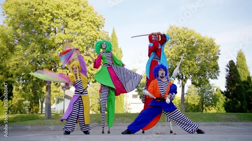Group of street performers in colorful costumes juggle and pose on stilts in a park, captured in slow motion