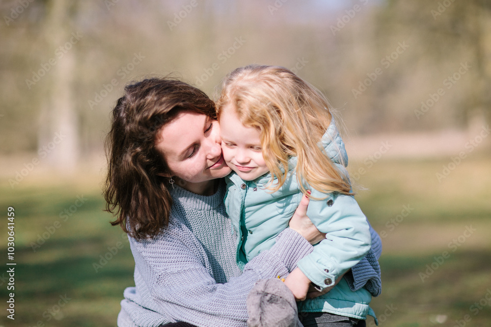Young beautiful mother and an adorable blond little daughter are hugging and kiss each other in a park, happy family Young beautiful mother and an adorable blond little daughter are hugging and kiss each other in a park, happy family