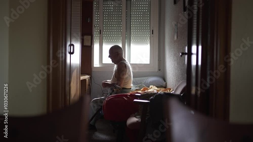 video of elderly man in his room in the morning, single bed and natural light. He gets up and walks with two walking sticks.