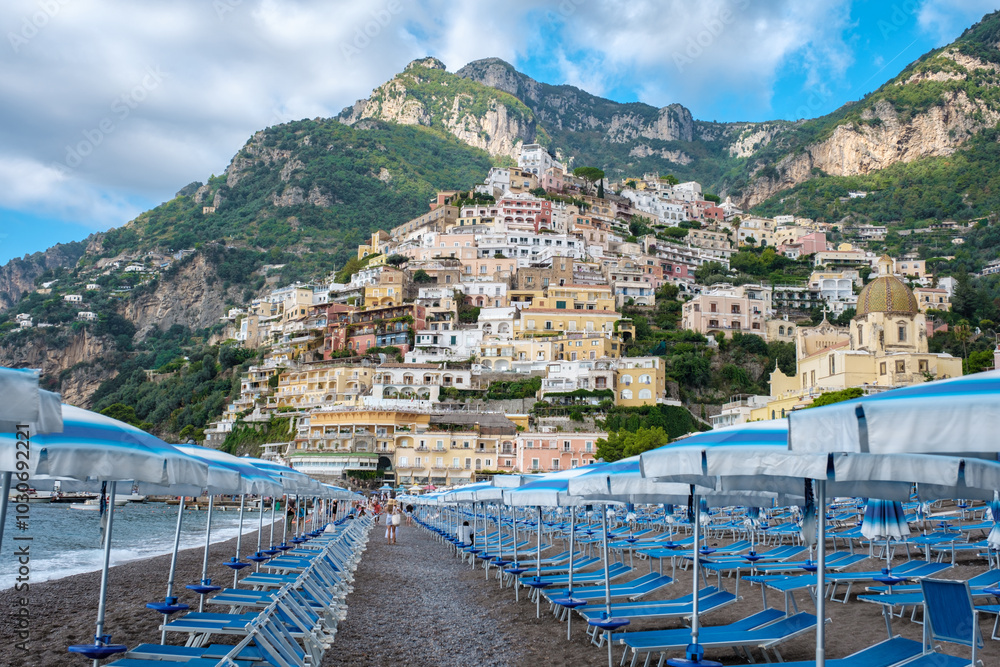 Naklejka premium Colorful buildings cascade down the hillside as visitors bask on Positano's sun-soaked shores, where iconic beach umbrellas dot the landscape beneath stunning cliffs.
