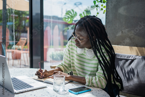 Young professional is working remotely at a cafe, taking notes in a notebook while using a laptop. She is wearing casual clothing and has headphones around her neck photo