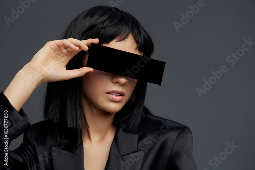 Woman in black suit holding a black box over her eyes, looking straight at the camera with a blank expression, highlighting a sense of mystery or concealment photo