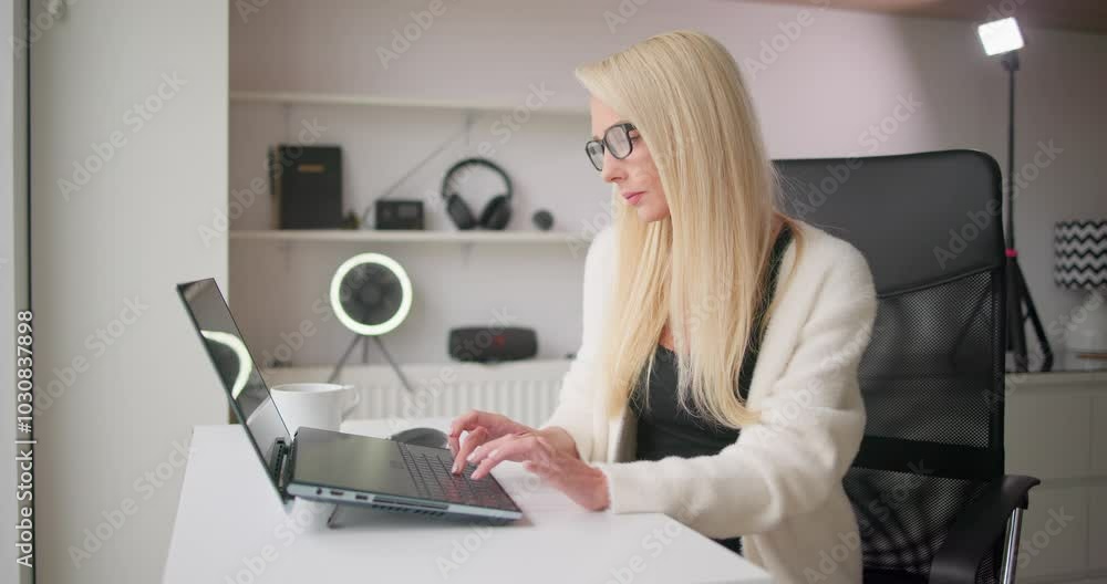 Young beautiful white woman blonde in casual clothes and glasses working or studying on laptop from home or office typing and looking at screen sitting in chair at table Young beautiful white woman blonde in casual clothes and glasses working or studying on laptop from home or office typing and looking at screen sitting in chair at table