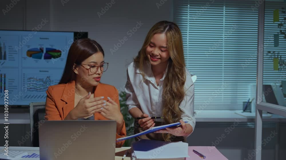 Two businesswomen work late in an office, discussing paperwork and collaborating on a laptop Two businesswomen work late in an office, discussing paperwork and collaborating on a laptop