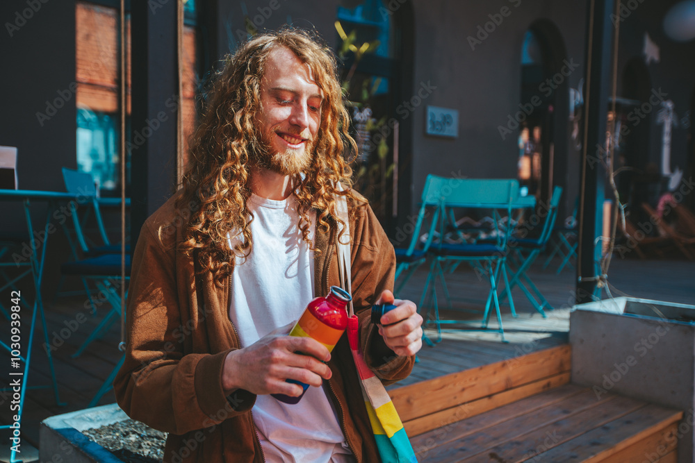 Young man with long hair takes a refreshing break at a cozy cafe, opening a vibrant reusable water bottle on a sunny day, embracing sustainability