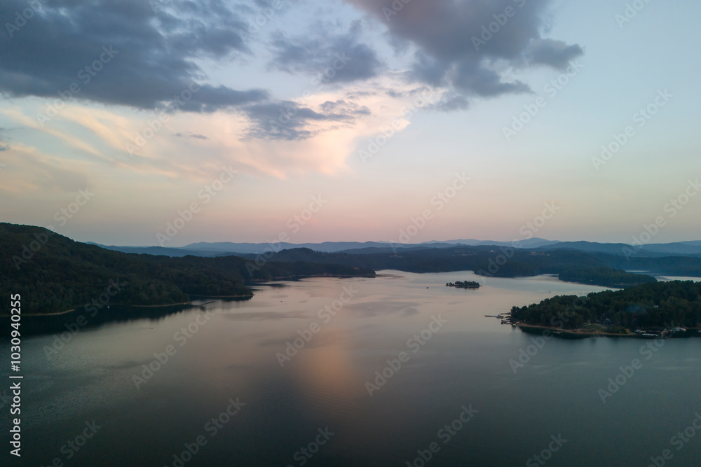 Fototapeta premium Aerial view of a tranquil lake surrounded by lush hills at sunset, showcasing beautiful reflections on the water's surface