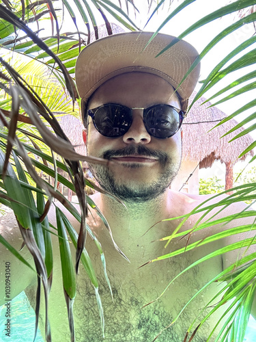 A person wearing sunglasses and a cap smiles for a selfie, surrounded by lush green palm leaves. The tropical setting features natural light and hints of a pool or hut in the background.