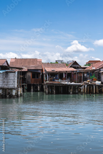 Photograph of traditional floating houses in George Town, Malaysia, captured on a bright, clear day with a vibrant blue sky. The serene scene showcases the unique waterfront architecture.