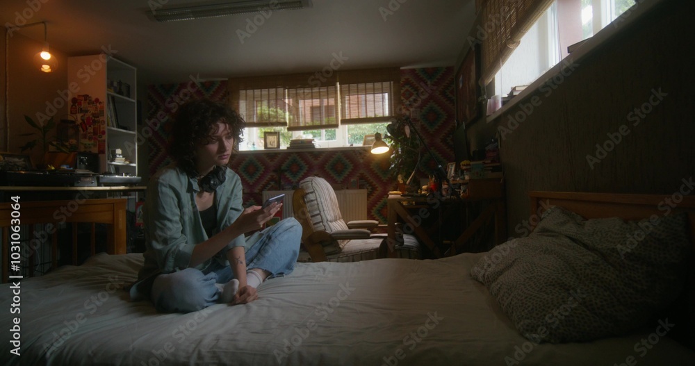 A young woman uses a mobile phone while sitting on a bed at home with a depressed look A young woman uses a mobile phone while sitting on a bed at home with a depressed look
