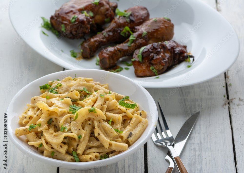 Cheese macaroni with herbs in a white ceramic bowl served with sticky grilled chicken white plate on a white wooden table Cheese macaroni with herbs in a white ceramic bowl served with sticky grilled chicken white plate on a white wooden table