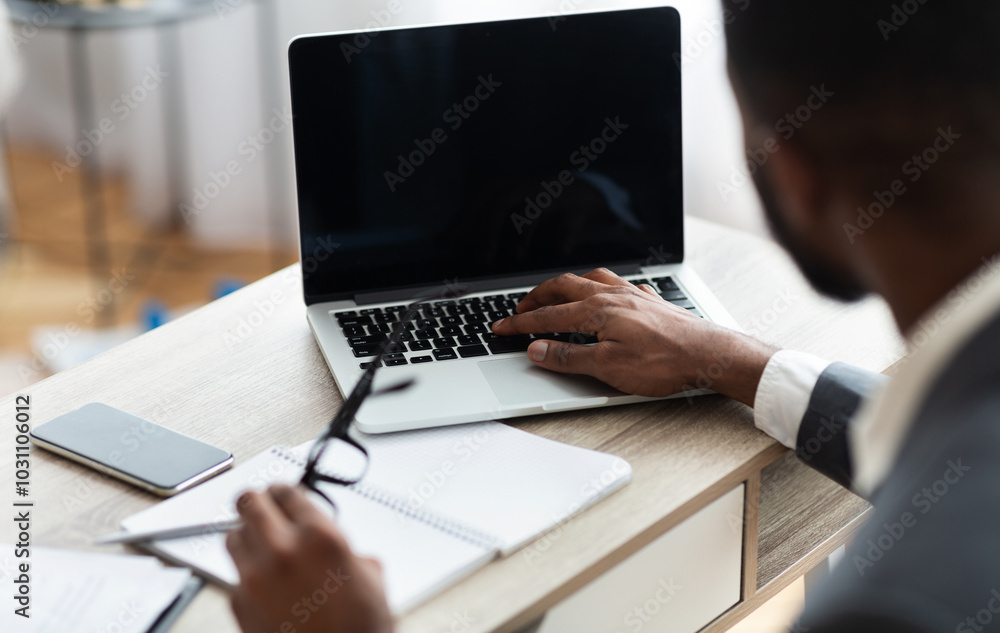 Unrecognizable black businessman working on laptop with blank screen on his workplace in modern office interior. Over shoulder shot, selective focus on device Unrecognizable black businessman working on laptop with blank screen on his workplace in modern office interior. Over shoulder shot, selective focus on device
