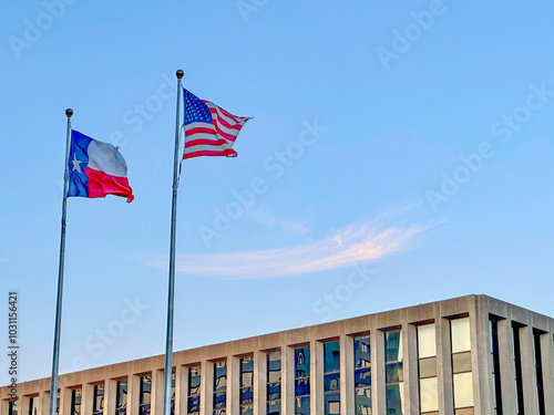 American flag and Texas flag blowing in the wind against a deep blue sky with concrete building in the background.