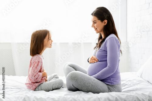 Pregnant mother and little daughter in pajamas touching bellies, sitting on bed face to face at home