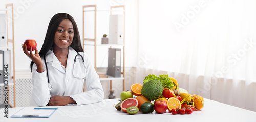 African american smiling female doctor dietologist sitting at workdesk, holding apple in her hand, empty space photo