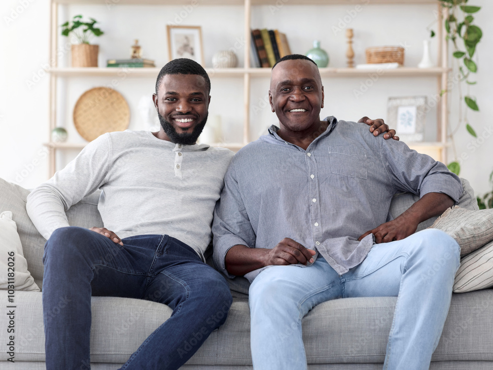 Portrait of adult black son and his senior father sitting on couch at home, posing to camera, free space Portrait of adult black son and his senior father sitting on couch at home, posing to camera, free space