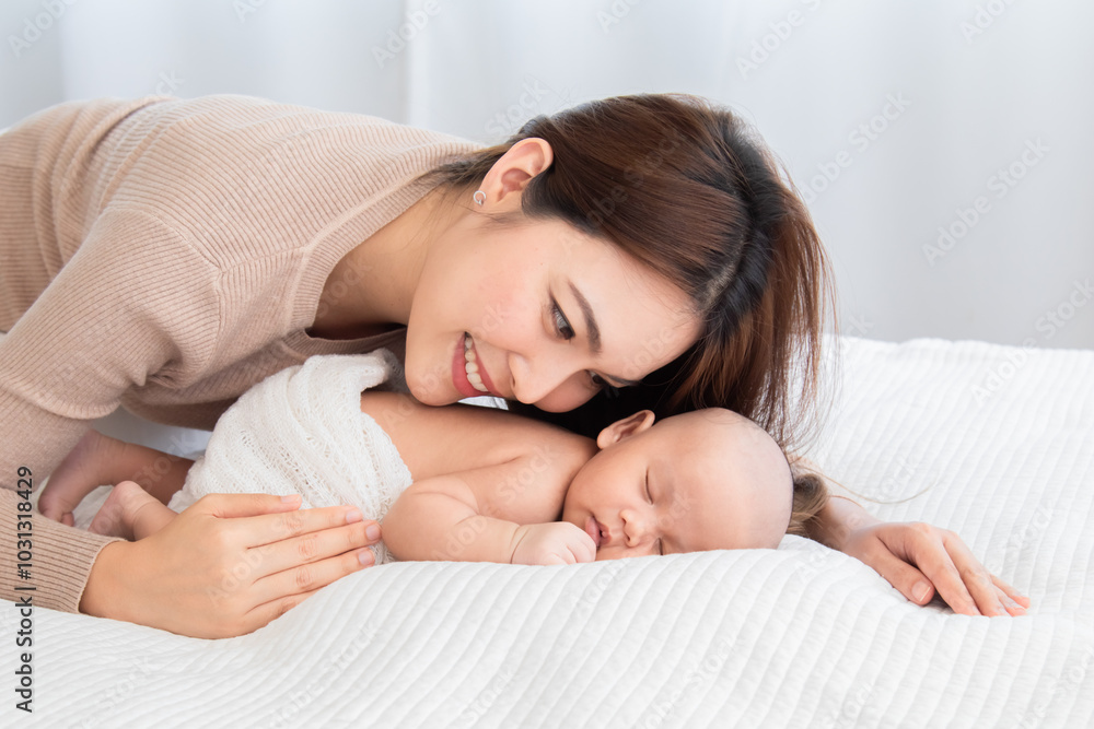 Asian young mother holding tenderly cuddles newborn baby in arms on to chest. Mother looking adorable infant with love and care. Woman and small child lying on bed together at home Asian young mother holding tenderly cuddles newborn baby in arms on to chest. Mother looking adorable infant with love and care. Woman and small child lying on bed together at home