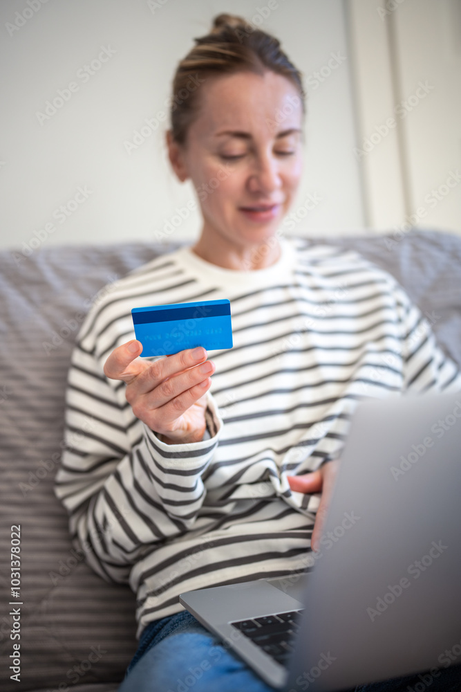 Convenience online shopping from home. Middle aged woman pays for internet purchases with credit card using laptop sitting on couch. Easy-to-use banking app for comfortable payment of utility bills. Convenience online shopping from home. Middle aged woman pays for internet purchases with credit card using laptop sitting on couch. Easy-to-use banking app for comfortable payment of utility bills.