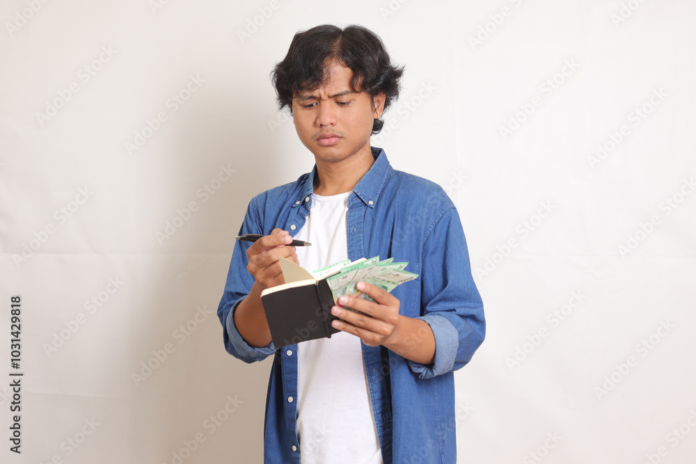 Portrait of attractive Asian man in blue shirt writing notes about personal finance while holding rupiah money banknote. Financial education concept. Isolated images on white background Portrait of attractive Asian man in blue shirt writing notes about personal finance while holding rupiah money banknote. Financial education concept. Isolated images on white background
