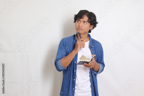 Portrait of confused Asian man in blue shirt writing on his notes while thinking about idea. Isolated images on white background photo