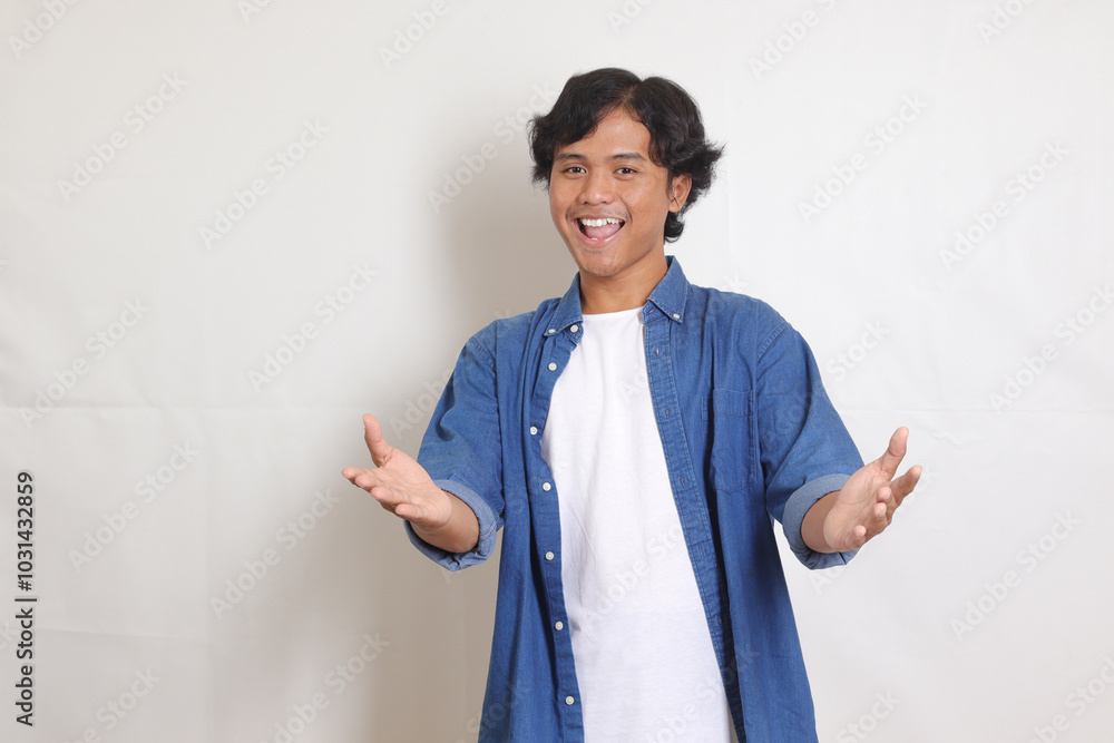 Portrait of excited Asian man in blue shirt spreading his hands sideways and holding two things, demonstrate products, welcoming someone. Isolated image on white background Portrait of excited Asian man in blue shirt spreading his hands sideways and holding two things, demonstrate products, welcoming someone. Isolated image on white background
