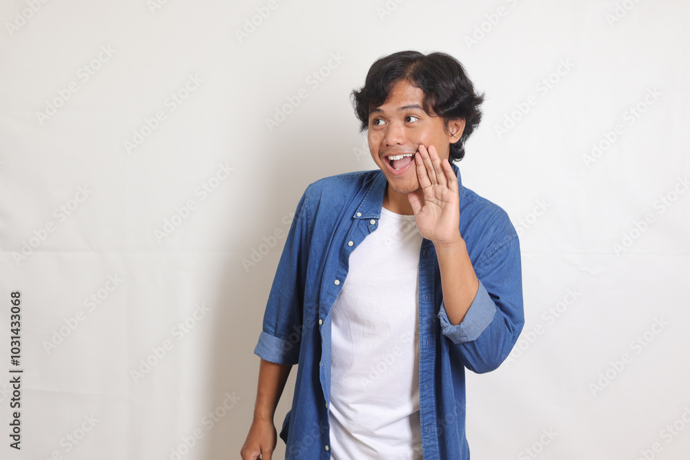 Portrait of excited Asian man in blue shirt whispering malicious talk conversation, hand on mouth telling secret rumor. Isolated image on white background Portrait of excited Asian man in blue shirt whispering malicious talk conversation, hand on mouth telling secret rumor. Isolated image on white background