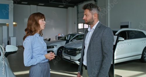 A car dealership manager in formal clothes smiles and talks to a customer standing near cars in a car dealership