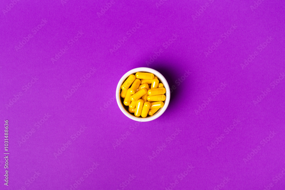 Flat lay view of dietary supplements and vitamins pills to support immune system and health. Meds to avoid illnesses and cold. Yellow vitamin capsules in a glass bowl from above on violet background. Flat lay view of dietary supplements and vitamins pills to support immune system and health. Meds to avoid illnesses and cold. Yellow vitamin capsules in a glass bowl from above on violet background.