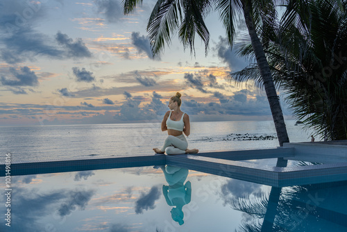 Young woman in a white clothes doing yoga on the beach near  swimming pool early morning in the summer. Sunrise sky in the background .