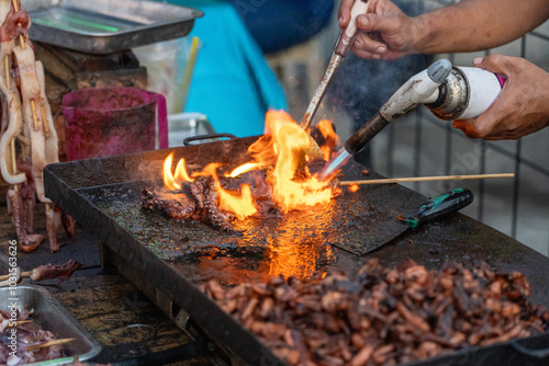 Street food vendor grilling octopus on kebab stick skewer using gas fire from flame torch - in Jalan Jogokariyan Mosque Ramadan Night market, Yogyakarta, for iftar (evening meal to break fast).