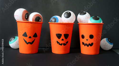 Halloween set of three orange jack o lantern glasses. Different facial expressions. Glasses are filled with round painted eyes of different colors. The eyes are also near the glasses. Black background photo