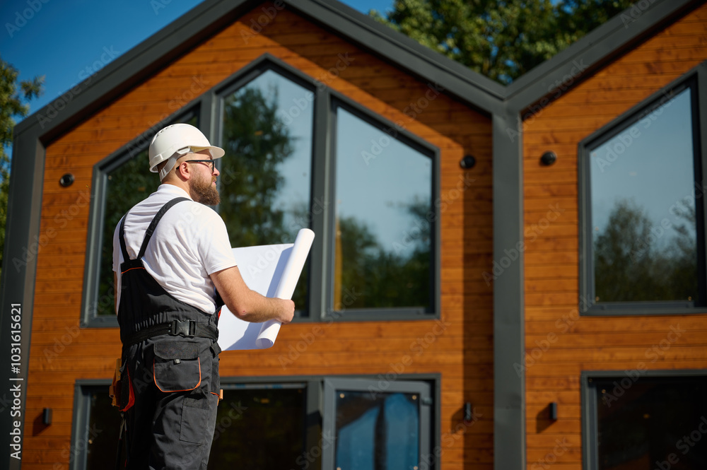 Architect hold plan. Bearded architect wearing hardhat holding project blueprints paper plans standing near comlex of new modern houses looking to something at construction site. Architect hold plan. Bearded architect wearing hardhat holding project blueprints paper plans standing near comlex of new modern houses looking to something at construction site.