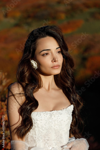 A woman in a white wedding dress stands on a hill before a forest