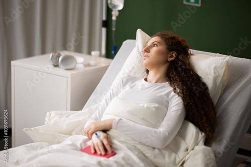 Resting in the hospital room, the young patient, with an oxygen tube in her nose, holds a phone, gazing out the window in quiet reflection.