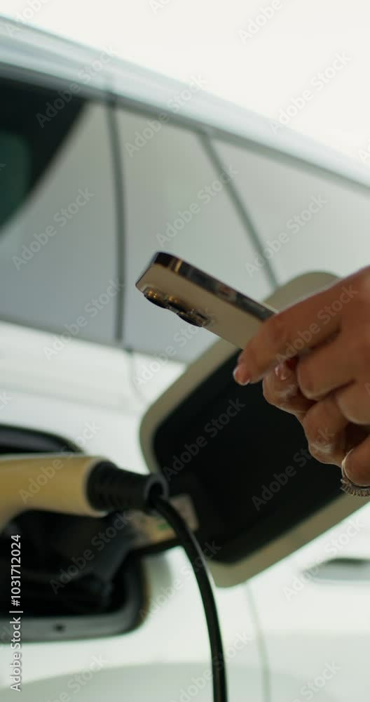 A woman uses a mobile phone standing near an electric car. Close-up of her hands. Charging cable in the connector A woman uses a mobile phone standing near an electric car. Close-up of her hands. Charging cable in the connector
