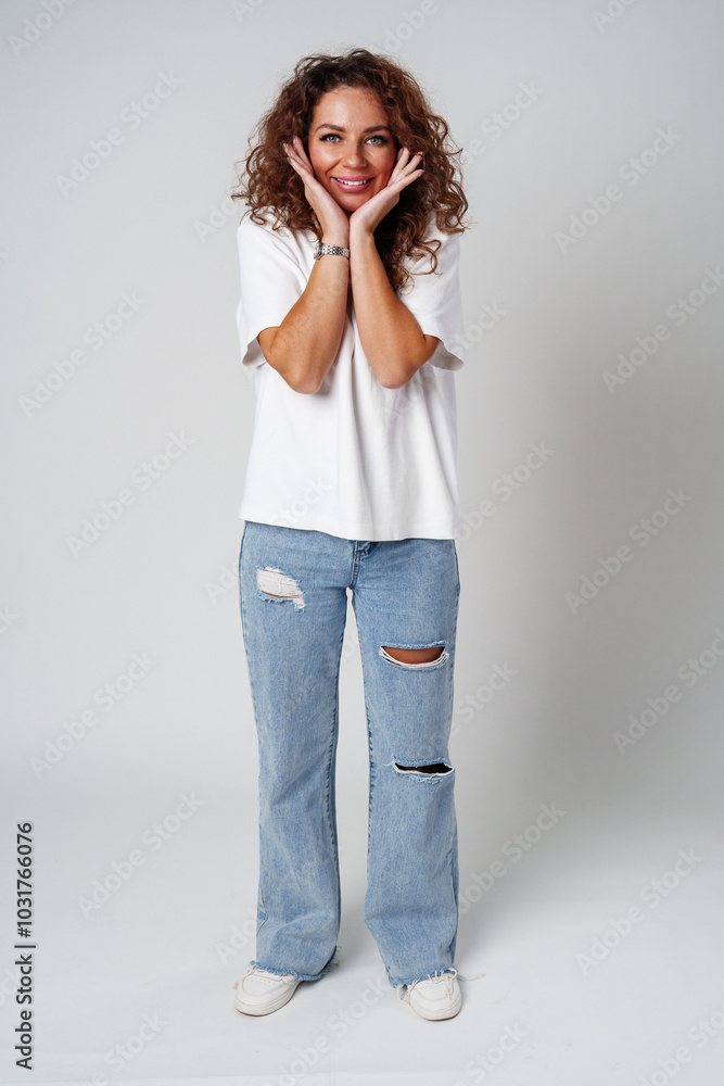 Woman with curly hair smiles joyfully while posing with hands on her cheeks against a light background Woman with curly hair smiles joyfully while posing with hands on her cheeks against a light background