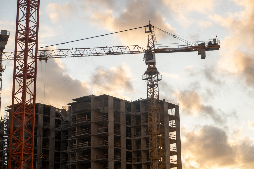 Cranes are lifting materials at a busy construction site as the sun sets, casting a warm glow over the ongoing building project Cranes are lifting materials at a busy construction site as the sun sets, casting a warm glow over the ongoing building project