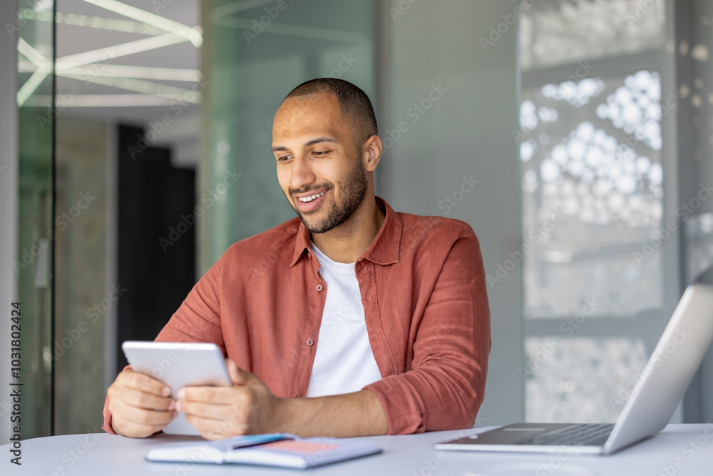 Smiling man using digital tablet in modern office setting. Engages in digital tasks with tablet and laptop. Casual attire suggests relaxed yet professional atmosphere. Smiling man using digital tablet in modern office setting. Engages in digital tasks with tablet and laptop. Casual attire suggests relaxed yet professional atmosphere.
