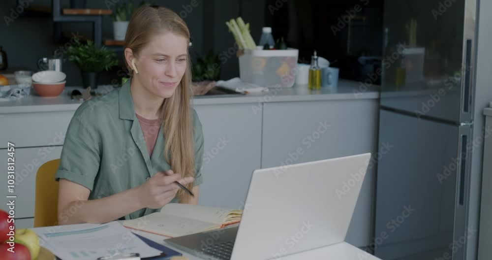 Female entrepreneur discussing business with partner during online video call speaking and writing in notebook using laptop and earphones in kitchen at home Female entrepreneur discussing business with partner during online video call speaking and writing in notebook using laptop and earphones in kitchen at home