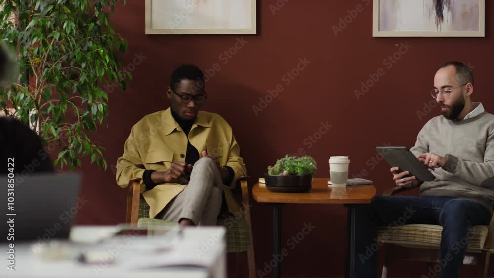 Girl working on laptop while two multi-ethnic young men sitting in armchairs and chatting during workday in office, Caucasian manager holding digital tablet Girl working on laptop while two multi-ethnic young men sitting in armchairs and chatting during workday in office, Caucasian manager holding digital tablet