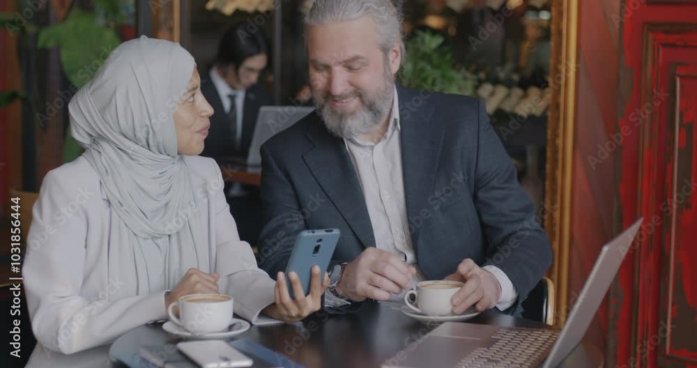 Young Middle Eastern businesswoman in hijab using smartphone swiping screen and talking to colleague mature Caucasian man at lunch break in cafe. Gadget and business concept. Young Middle Eastern businesswoman in hijab using smartphone swiping screen and talking to colleague mature Caucasian man at lunch break in cafe. Gadget and business concept.