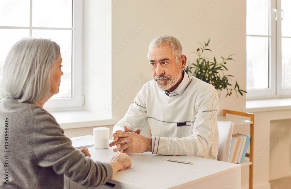 Obraz premium Elderly husband and wife, sitting together at a table at home, engage in a serious conversation. The senior couple talking about important matters, showing strong bond and communication as a couple.