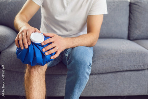 Photos Unrecognizable man with a blue ice pack on his injured knee sitting on a gray couch at home